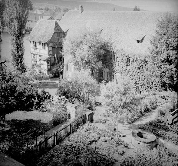 View from high-up of a garden and monastery buildings. There are plain tiles on the roofs. The walls are ivy-clad. Photo in black and white.